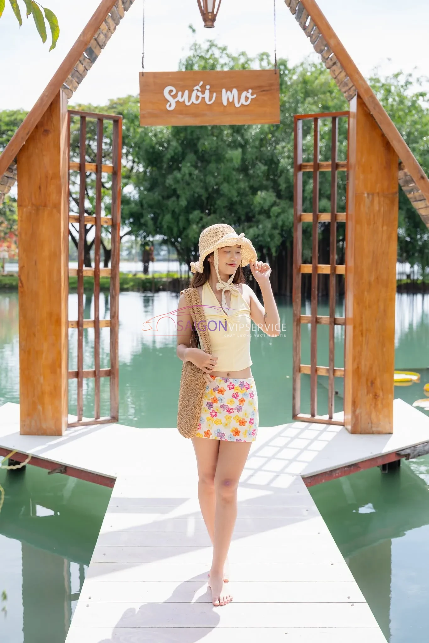 Du khách nữ đội mũ rộng vành tạo dáng chụp ảnh check-in tại tiểu cảnh bằng gỗ bên hồ nước ở Khu du lịch Suối Mơ